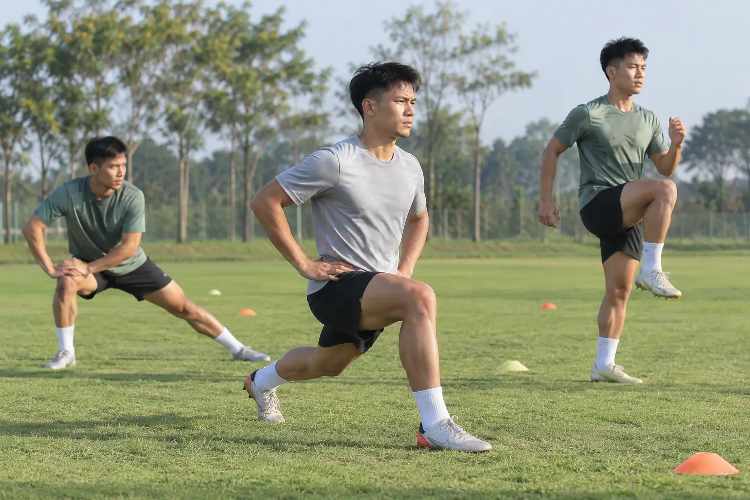 Ba cầu thủ bóng đá thực hiện động tác chùng chân, giãn cơ ngang và nâng cao gối trên sân cỏ dưới ánh nắng buổi sáng.