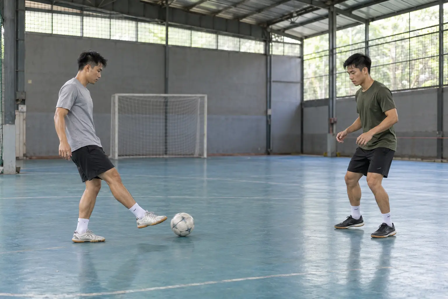 Hai người tập bóng đá phối hợp chuyền và nhận bóng trên sân trong nhà có mái che.