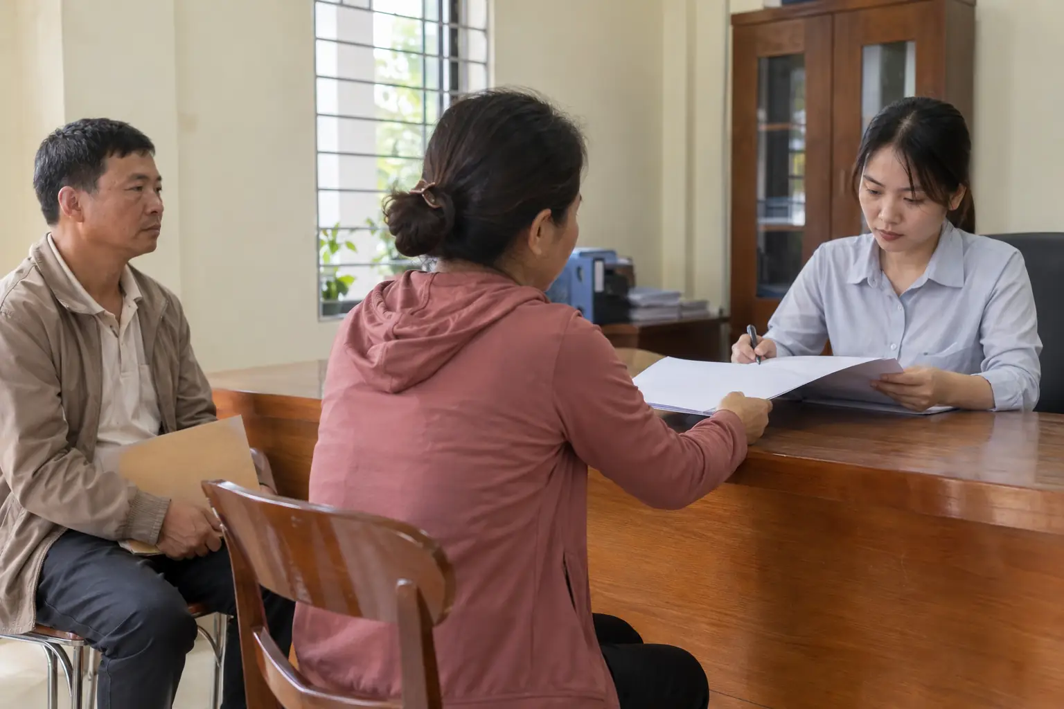 Người phụ nữ đưa hồ sơ cho nhân viên kiểm tra tại bàn tiếp nhận, bên cạnh là một người đàn ông ngồi chờ cầm phong bì.