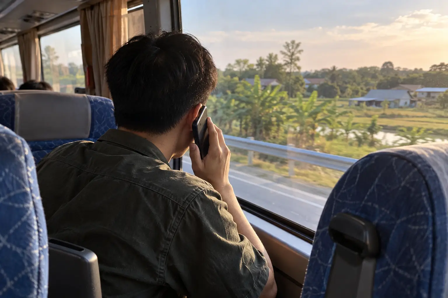 Nam hành khách nhìn từ phía sau, áp điện thoại vào tai khi ngồi cạnh cửa sổ xe khách, bên ngoài là cảnh đồng quê.