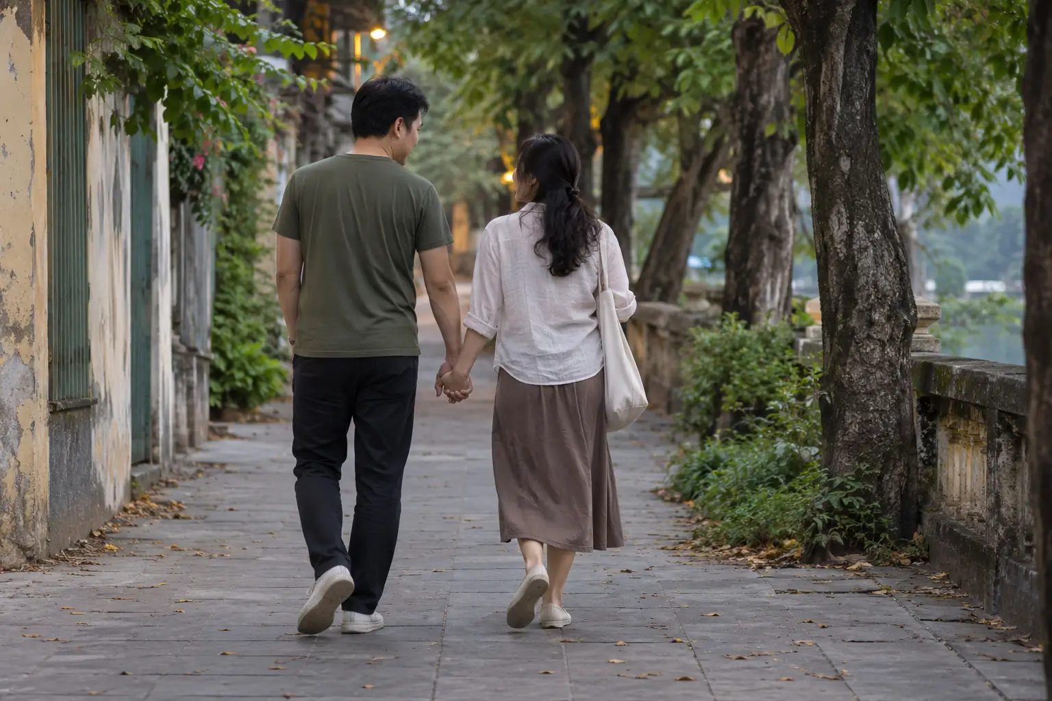 Một đôi nam nữ vô danh nắm tay đi trên lối lát đá dưới hàng cây, nhìn từ phía sau.