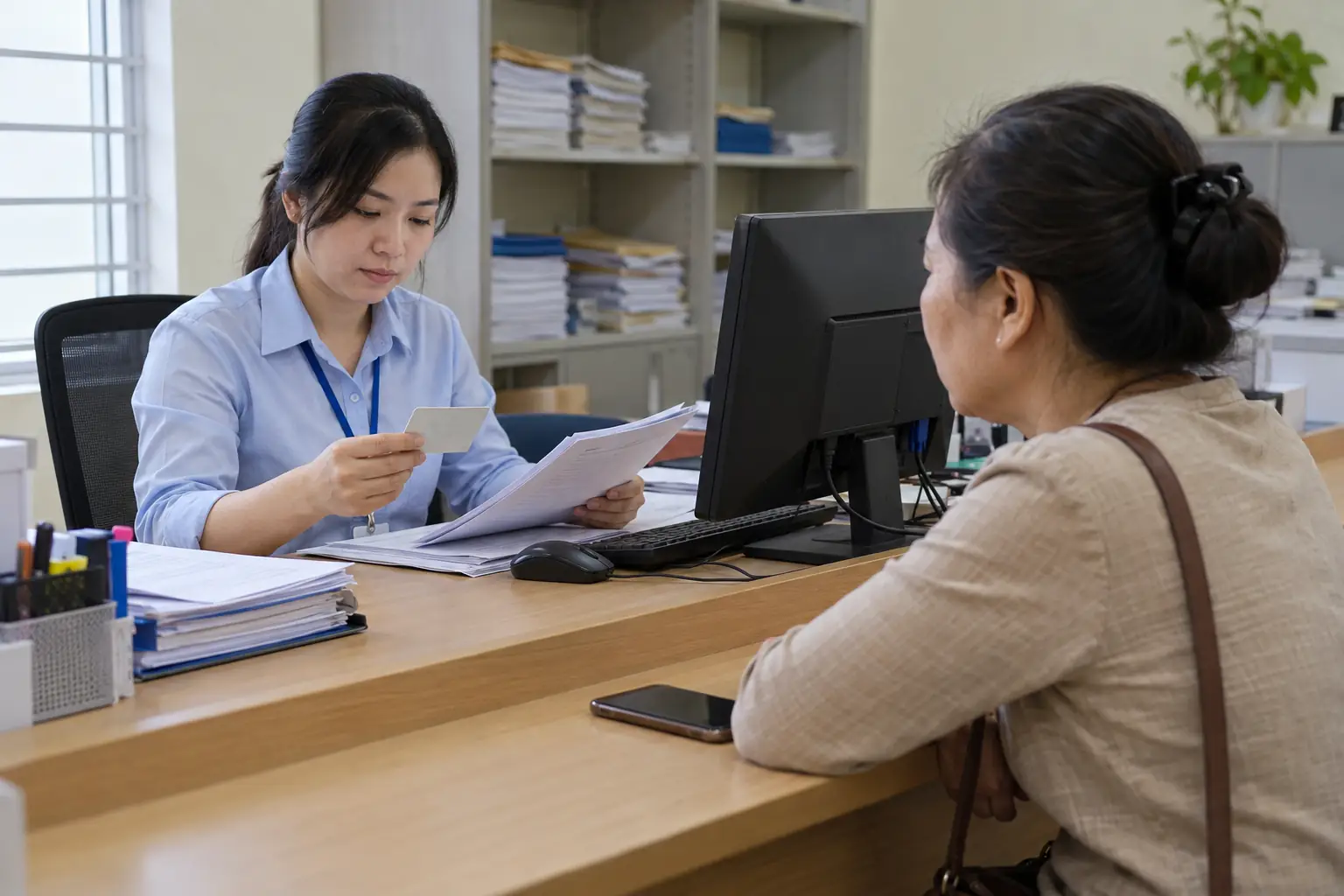 Nhân viên tại quầy đối chiếu thẻ và hồ sơ trước một phụ nữ đang ngồi chờ, bên cạnh là máy tính và điện thoại đặt trên bàn.