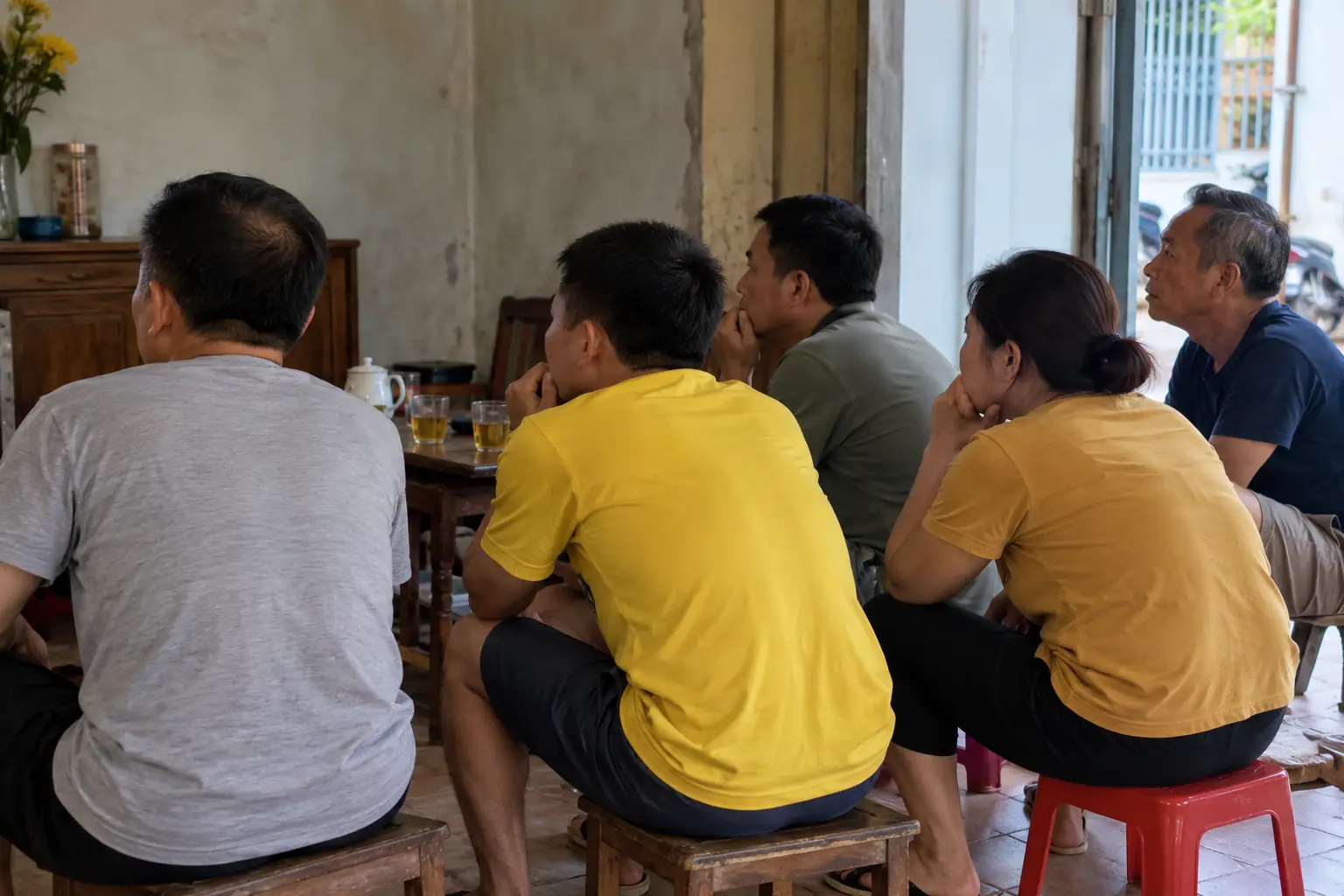 Năm người lớn ngồi sát nhau trong căn phòng giản dị, nhìn từ phía sau và cùng chăm chú hướng về phía ngoài khung hình.