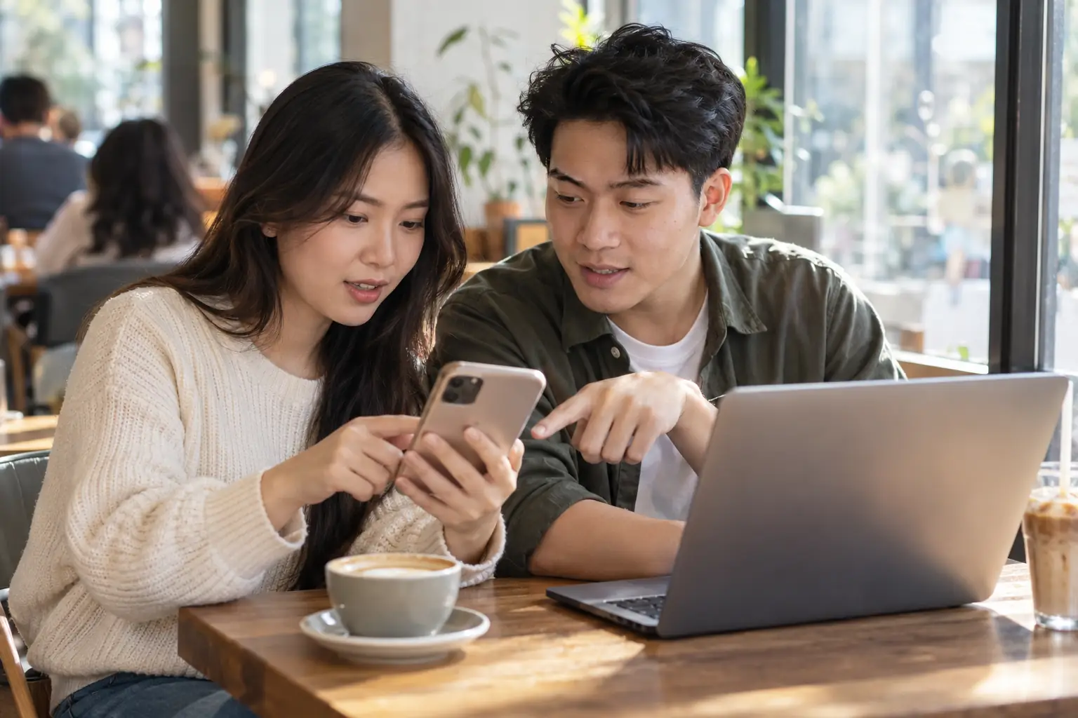 Hai người trẻ cùng xem điện thoại, người nam chỉ vào màn hình bên cạnh máy tính xách tay trên bàn cà phê.